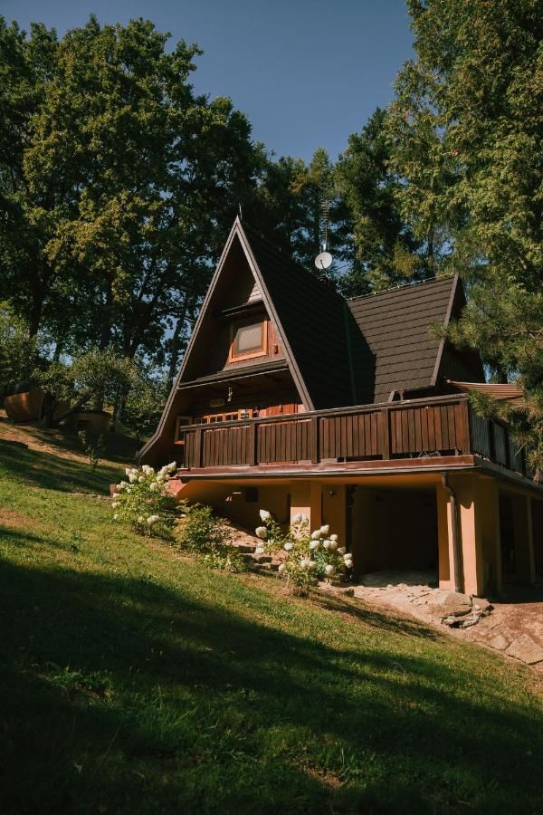 Drewniany dom z balkonem na trawiastym zboczu, wokół wysokie drzewa